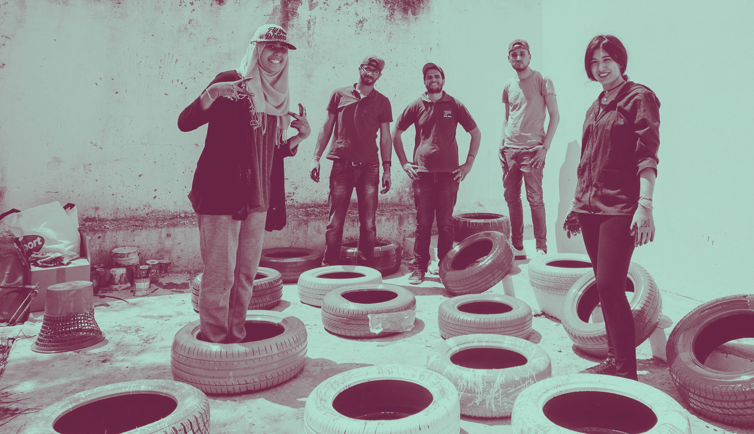 A group of young women and men stand surrounded by car tyres that have been painted in bright colours. They are smiling to camera.