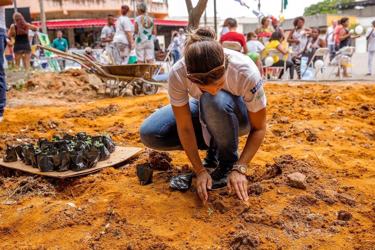 A woman in jeans and a white t-shirt kneels down to the ground to plant a young seedling. The soil is bright orange and looks freshly turned. A wheelbarrow sits in the background.
